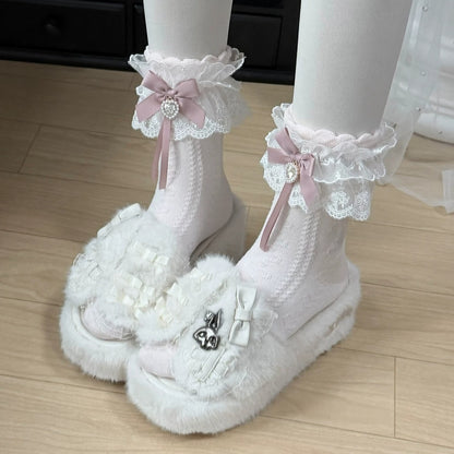 White lace ruffled socks with pink bows and white fluffy platform sandals with silver heart charms, shown on a person's feet on a light wood floor.