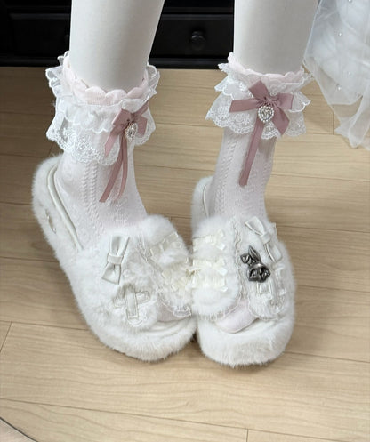 White fuzzy Mary Jane platform shoes with lace ruffles, pink ribbon bows, and decorative bows on the toes, shown on a mannequin's feet against a light wood floor.