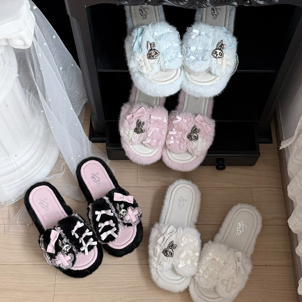 Four pairs of fluffy slide sandals displayed on a black rack and floor. The designs include light blue, pink, black and pink, and white versions, all decorated with bows, ribbons, and skull embellishments.