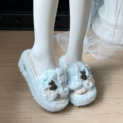A pair of fluffy light blue open-toe slippers with decorative bows and dark brown skull embellishments, shown on a white mannequin leg against a light wood floor.