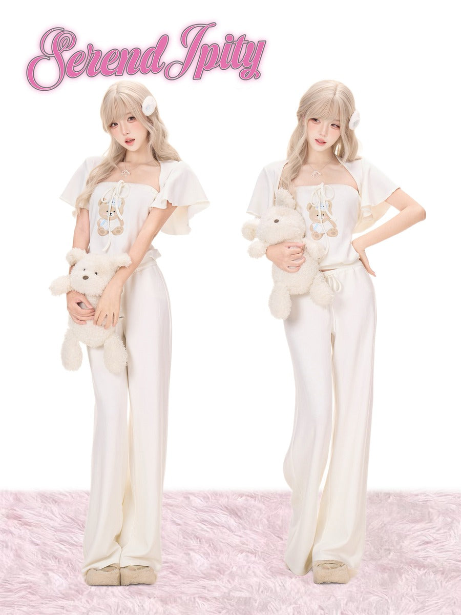 Two views of a model wearing a cream-colored two-piece outfit with ruffled sleeves and wide-leg pants, holding a plush teddy bear.