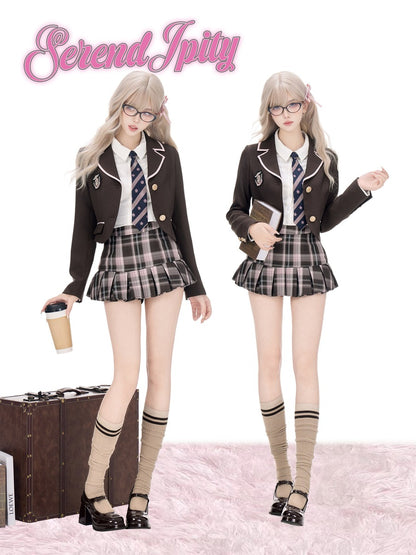 Two models wearing a school uniform cosplay set consisting of a dark brown blazer, white shirt, striped tie, plaid pleated skirt, and beige knee-high socks. The image features a pink 'Serendipity' logo at the top left and a brown vintage suitcase on the bottom left, set against a white background with a pink faux fur floor.