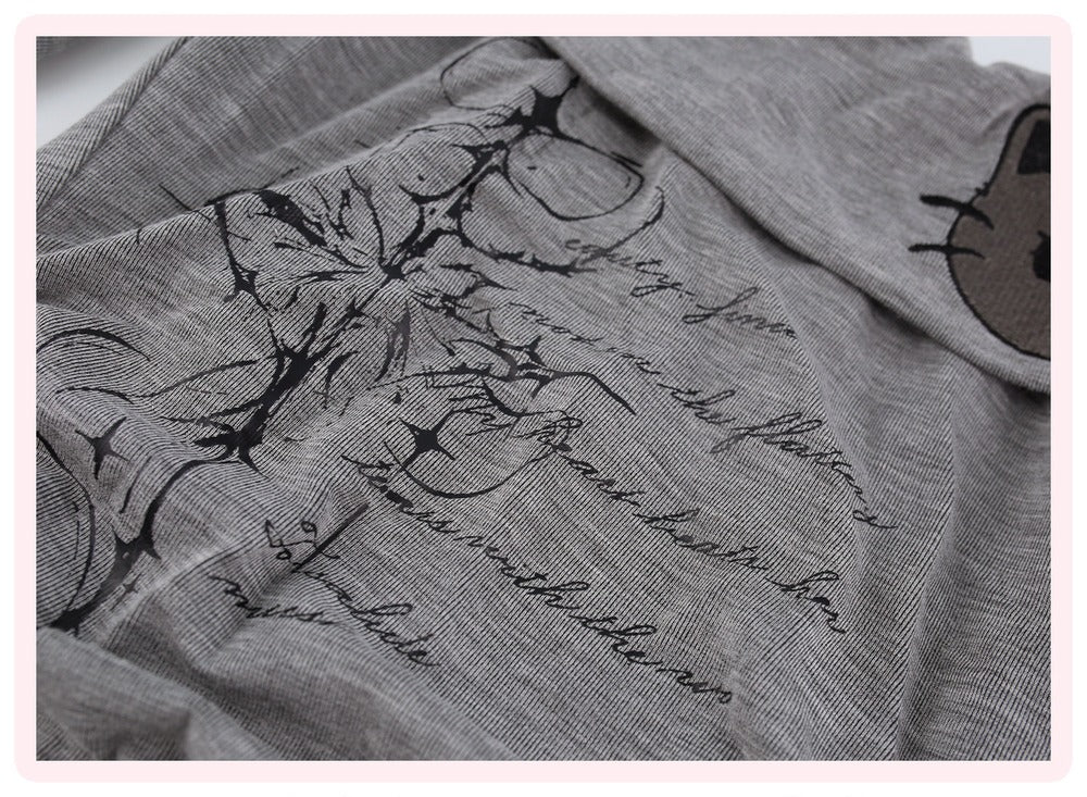 Close-up of a gray fabric garment with black floral line art, cursive script text, and a partial cartoon face patch on the sleeve.