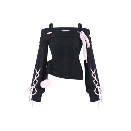 A black ribbed off-the-shoulder long-sleeve top with light pink accents, including side laces, a bow, and a pom-pom detail, shown against a plain white background.