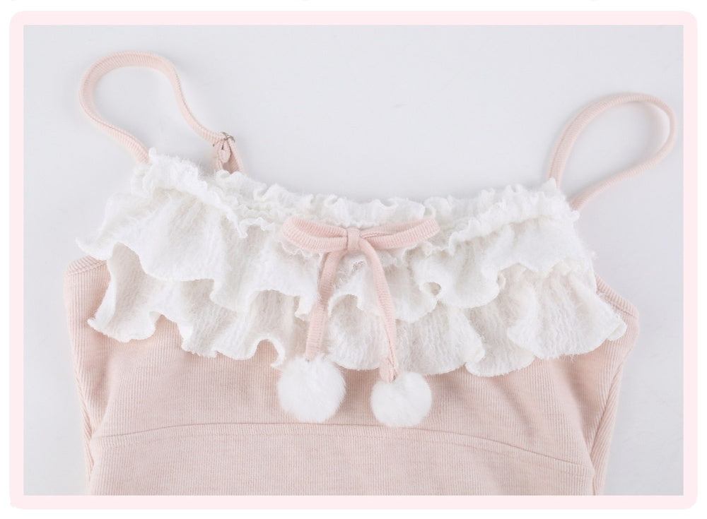 A pale pink ribbed knit camisole top featuring a white ruffled lace collar, a light pink bow, and two white pom-poms, shown against a light gray background with a pink border.
