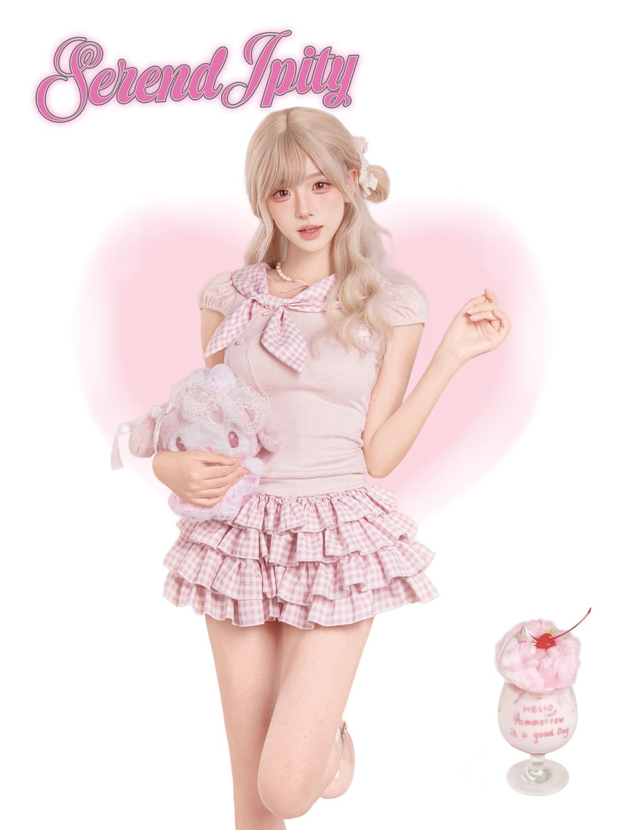 A young woman with blonde hair models a pink and white gingham outfit consisting of a short-sleeve top with a large bow and a ruffled mini skirt. She is holding a plush toy and standing in front of a pink heart-shaped glow on a white background