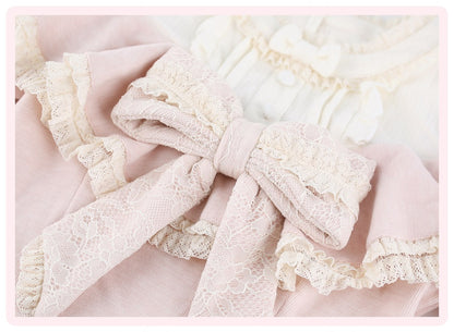 A close-up of a light pink garment with a large cream lace bow and matching ruffled trim, layered over a white ruffled fabric.