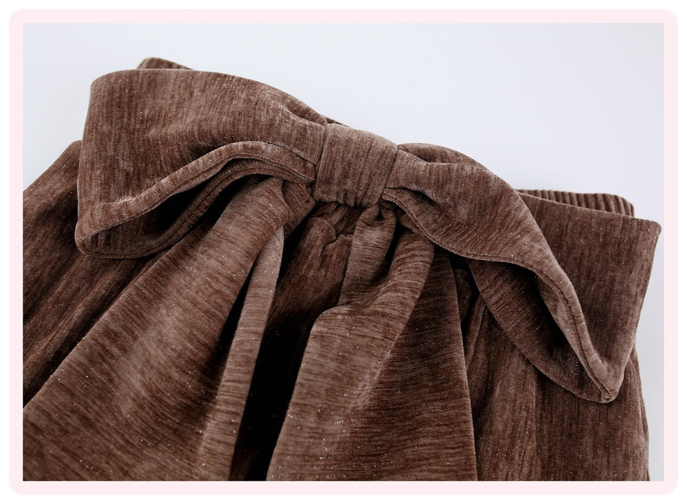 A close-up of a brown corduroy fabric accessory, likely a bag or pouch, featuring a large decorative bow on the front and gathered pleated fabric below, set against a plain light background.