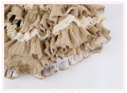 A close-up of a beige and cream ruffled textile with layered frills, lace trim, and a gathered ruffled edge, shown on a plain light background.