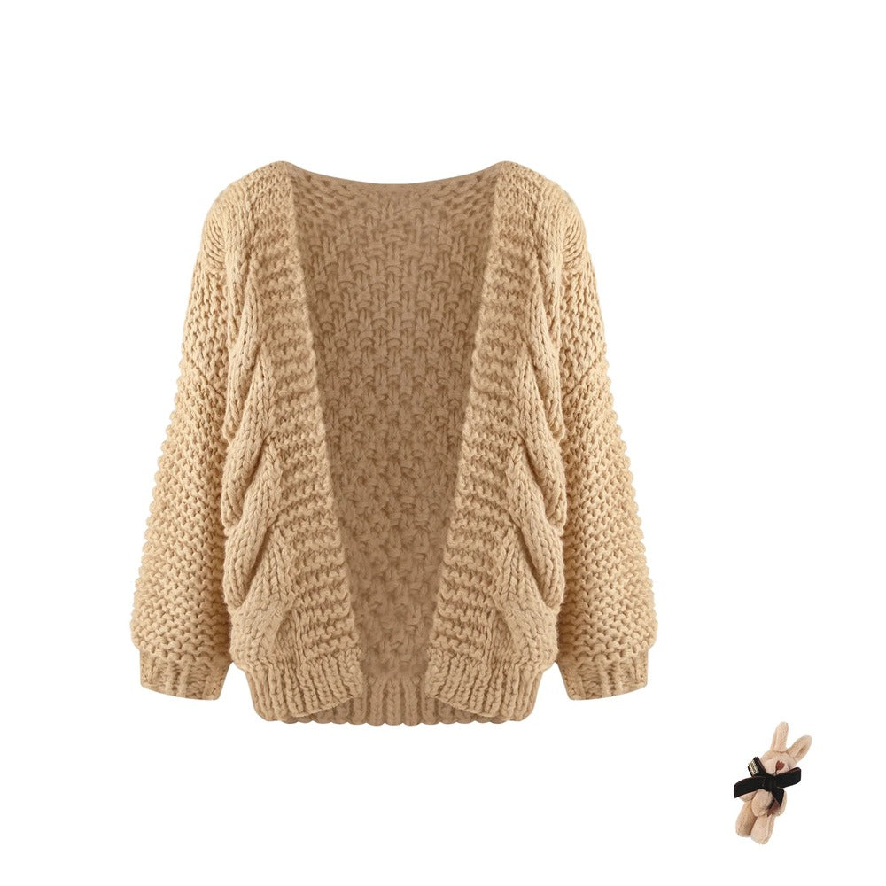 A beige chunky cable-knit open-front cardigan sweater with long sleeves, shown against a plain white background. A small plush bunny accessory is visible in the bottom right corner.