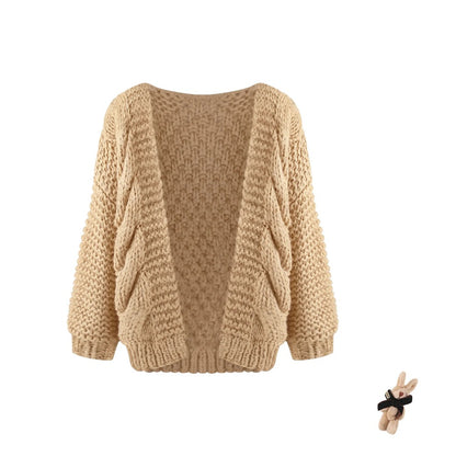 A beige chunky cable-knit open-front cardigan sweater with long sleeves, shown against a plain white background. A small plush bunny accessory is visible in the bottom right corner.