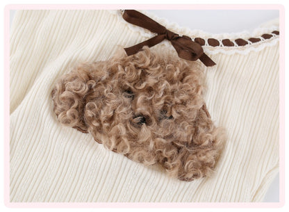 A close-up of a cream ribbed knit garment with a brown ribbon bow at the neckline and a textured brown curly hair accessory attached near the chest.