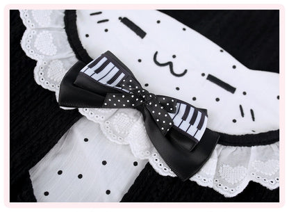 A close-up of a black and white cat-themed accessory, likely a headband or hair accessory, featuring a white fabric panel with black polka dots, embroidered cat face details, lace trim, and a large bow combining a black-and-white piano keyboard pattern with white polka dots.