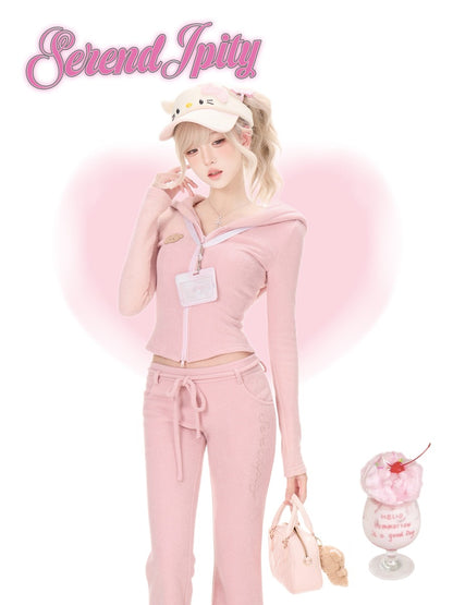 A model wearing a pink zip-up hoodie and matching pink sweatpants with a pink cat-themed cap, posing in front of a pink heart graphic on a white background