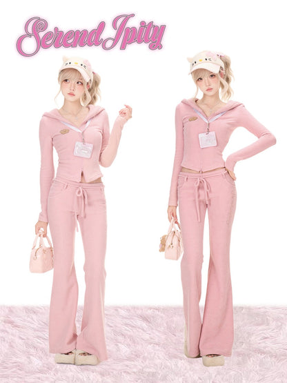 Two models wearing a pink two-piece tracksuit with a hooded long-sleeve top and matching wide-leg pants, styled with a white cat-ear hat and a small pink handbag against a white background with a pink fluffy rug.