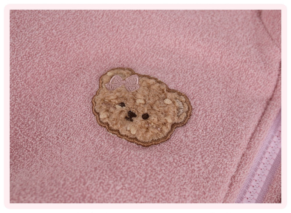 A close-up of a pink textured fabric featuring a teddy bear face applique patch with a small bow on the head.