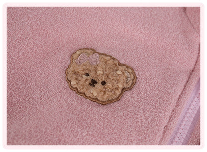 A close-up of a pink textured fabric featuring a teddy bear face applique patch with a small bow on the head.