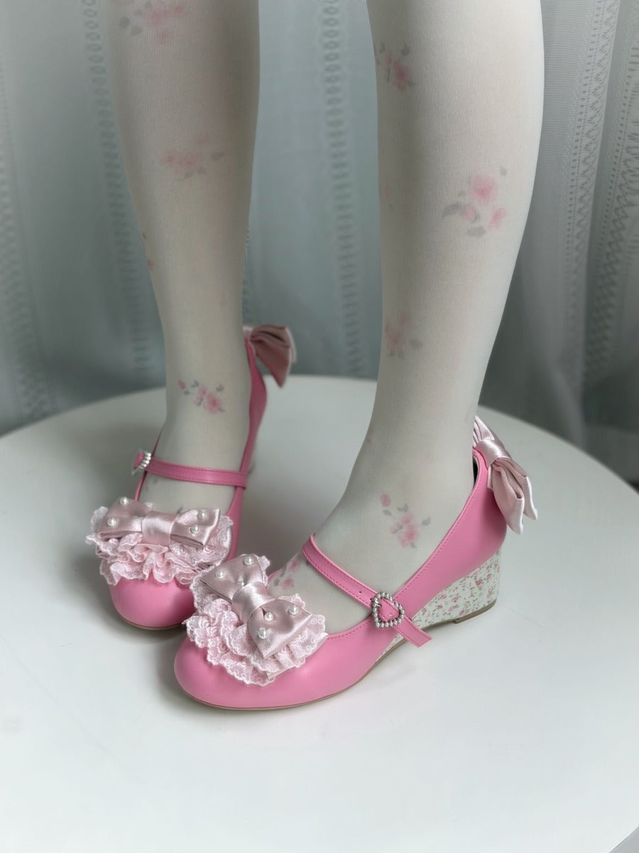 Pink Mary Jane style wedge shoes with ruffled bows and floral lace details, shown on a doll's feet wearing matching floral tights against a soft gray curtain background.
