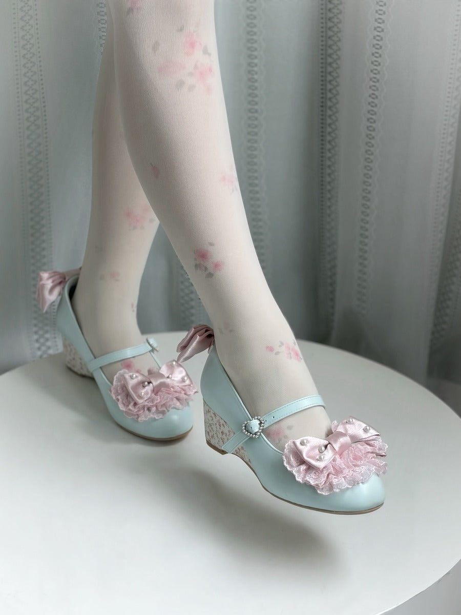 A pair of pastel light blue Mary Jane-style shoes with pink fabric roses and bows, worn with sheer white tights featuring a pink floral print, shown on a white table against a gray curtain background.