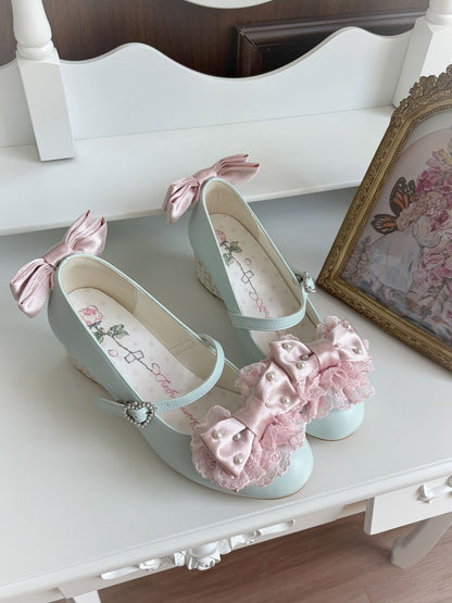 A pair of pastel mint green Mary Jane-style shoes with pink satin bows and ruffled trim, displayed on a white wooden surface next to a framed picture.