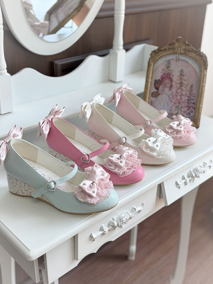 Three Mary Jane style shoes with wedge heels are arranged on a white vanity table. The shoes are light blue, pink, and cream, each decorated with a lace flower, a bow, and a small rhinestone buckle.