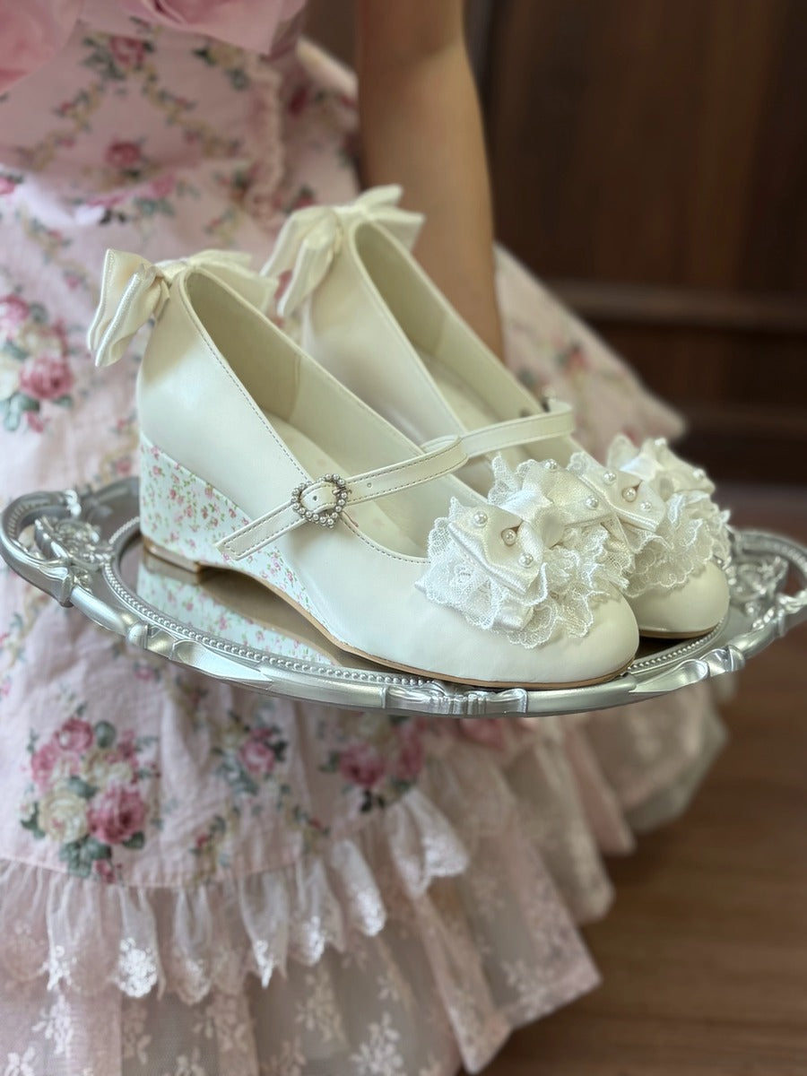 A pair of cream-colored Mary Jane style shoes with floral lace details and ribbon bows, displayed on a silver tray held by a person wearing a pink floral dress.