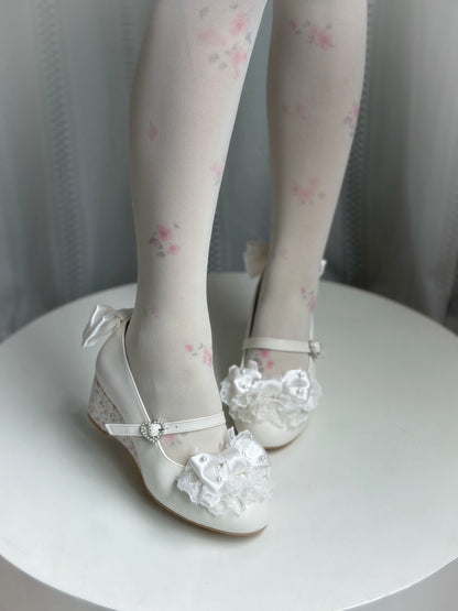 White Mary Jane low-heeled shoes with floral lace details, shown on a doll with white floral tights on a white round table.