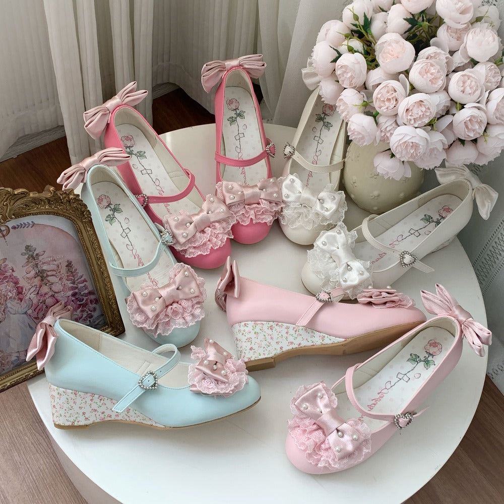 Six pastel wedge shoes arranged on a round white table, featuring pink, light blue, and white colors with floral lace-up bows, ruffled embellishments, and floral-patterned midsoles. The shoes are styled with a bouquet of pink roses, a gold picture frame, and soft curtains in the background.