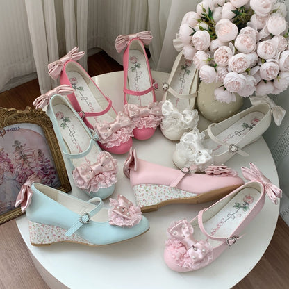 Six pastel wedge shoes arranged on a round white table, featuring pink, light blue, and white colors with floral lace-up bows, ruffled embellishments, and floral-patterned midsoles. The shoes are styled with a bouquet of pink roses, a gold picture frame, and soft curtains in the background.
