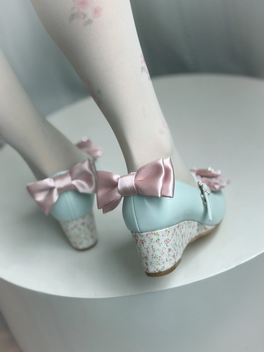 A pair of pastel light blue wedge shoes with large pink satin bows and floral-patterned heels, shown on a person's feet wearing white tights with a floral print, sitting on a white cylindrical pedestal.
