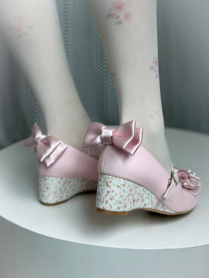 A pair of light pink Mary Jane-style block-heel shoes with large satin bows and floral wedge heels, shown on a model's feet wearing white floral tights.