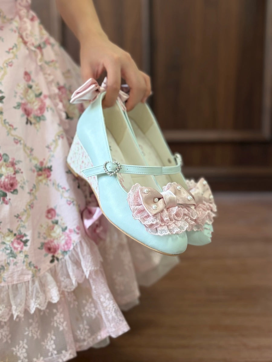 A person in a pink floral dress holds up a pair of pastel light blue Mary Jane shoes decorated with pink ruffles and bows, set against a warm wooden background.