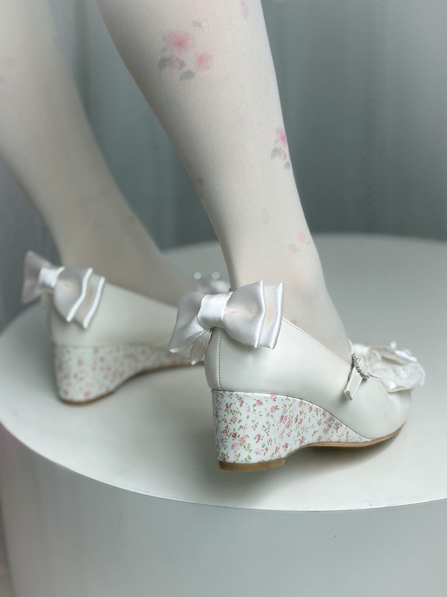 White Mary Jane wedge shoes with satin bows and a delicate pink floral print on the wedge and toe area, shown on a model wearing white tights with pink floral patterns.