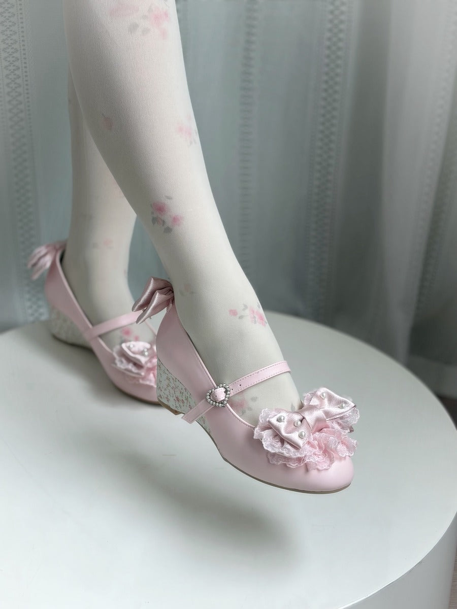 A pair of light pink Mary Jane-style shoes with ruffled floral embellishments and lace-up straps, shown on a model wearing white floral tights, sitting on a white round table.
