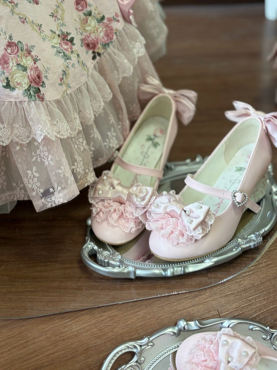A pair of pale pink Mary Jane shoes with ruffled bows and lace details, displayed on an ornate silver tray on a wooden floor next to a floral dress.