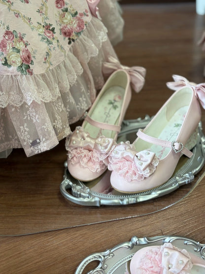 A pair of pale pink Mary Jane shoes with ruffled bows and lace details, displayed on an ornate silver tray on a wooden floor next to a floral dress.