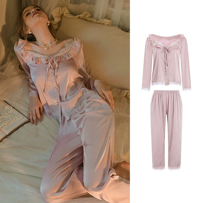 A women's two-piece pajama set in a soft light pink satin-like fabric, shown both worn by a woman reclining on a bed and as a clean product composite on a white background. The pajama set includes a long-sleeve top with ruffled trim and a tie at the neckline, matching drawstring pants, and lace trim at the cuffs.