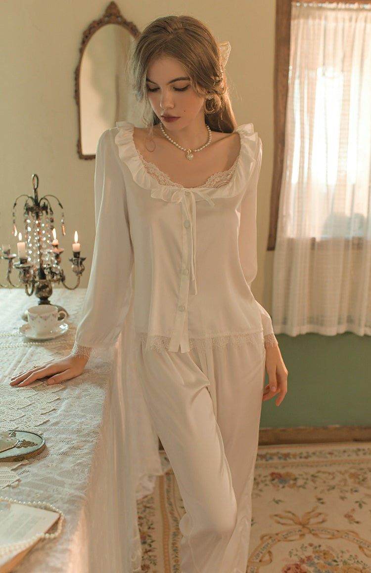 A woman wearing a white long-sleeve pajama set with ruffled trim and a tie-neck detail, standing in a softly lit vintage-style room beside a table with candles and a mirror.