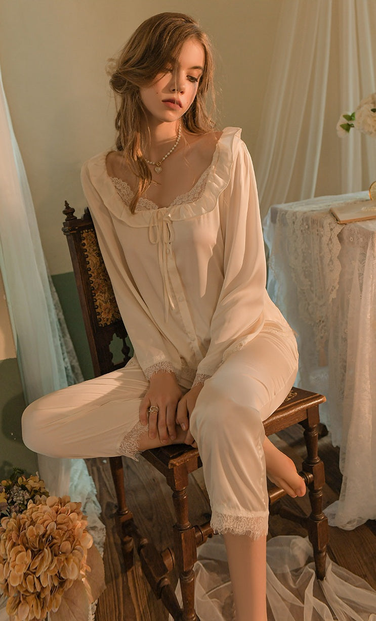 A woman wearing a cream-colored sheer lingerie set with lace trim, sitting on a wooden chair in a softly lit room with draped fabric and flowers.