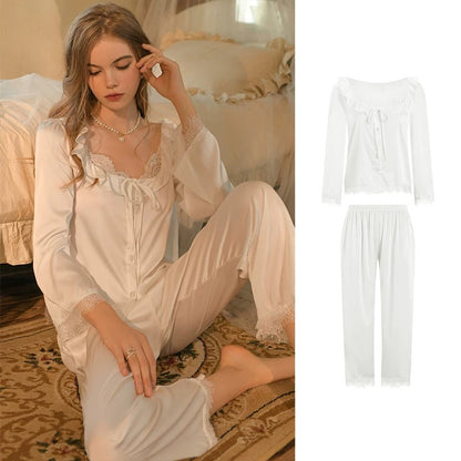 A woman wearing a two-piece white satin pajama set sits on a patterned rug in a softly lit bedroom. The set includes a long-sleeve top with lace trim and a tie at the neckline, plus matching loose pants.