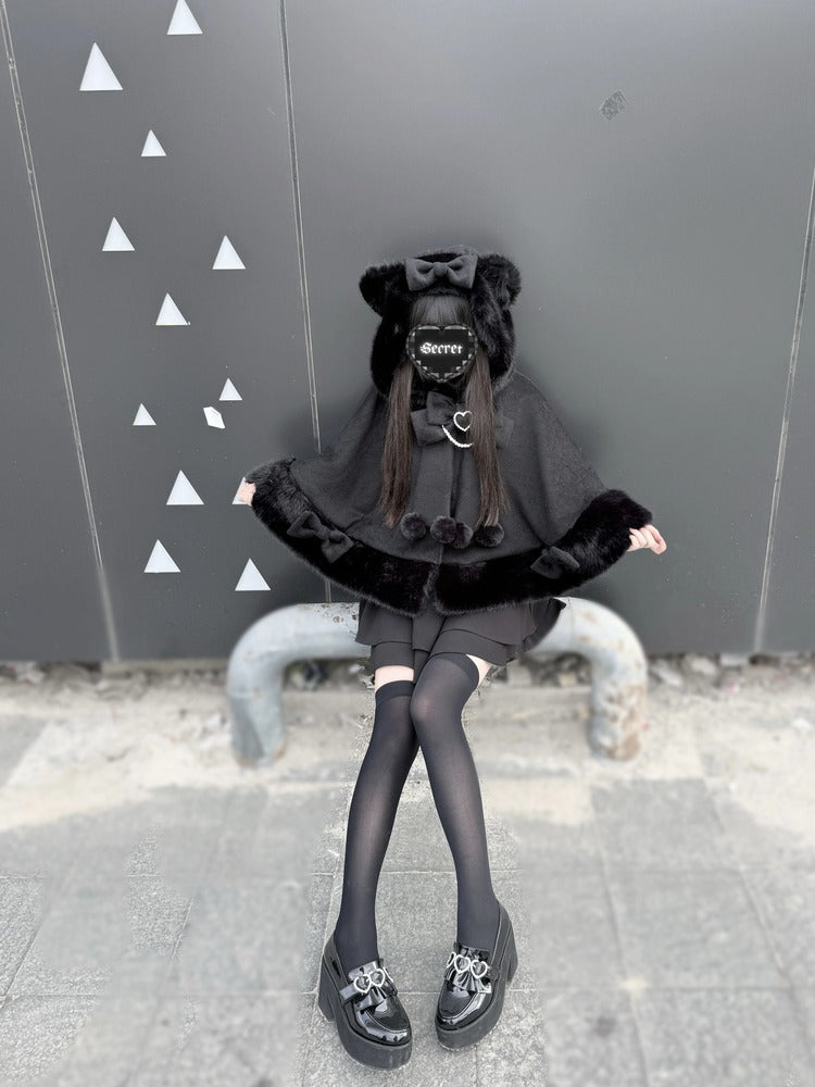 Person wearing a black outfit with a cape and shoes in front of a gray wall with white triangle patterns.