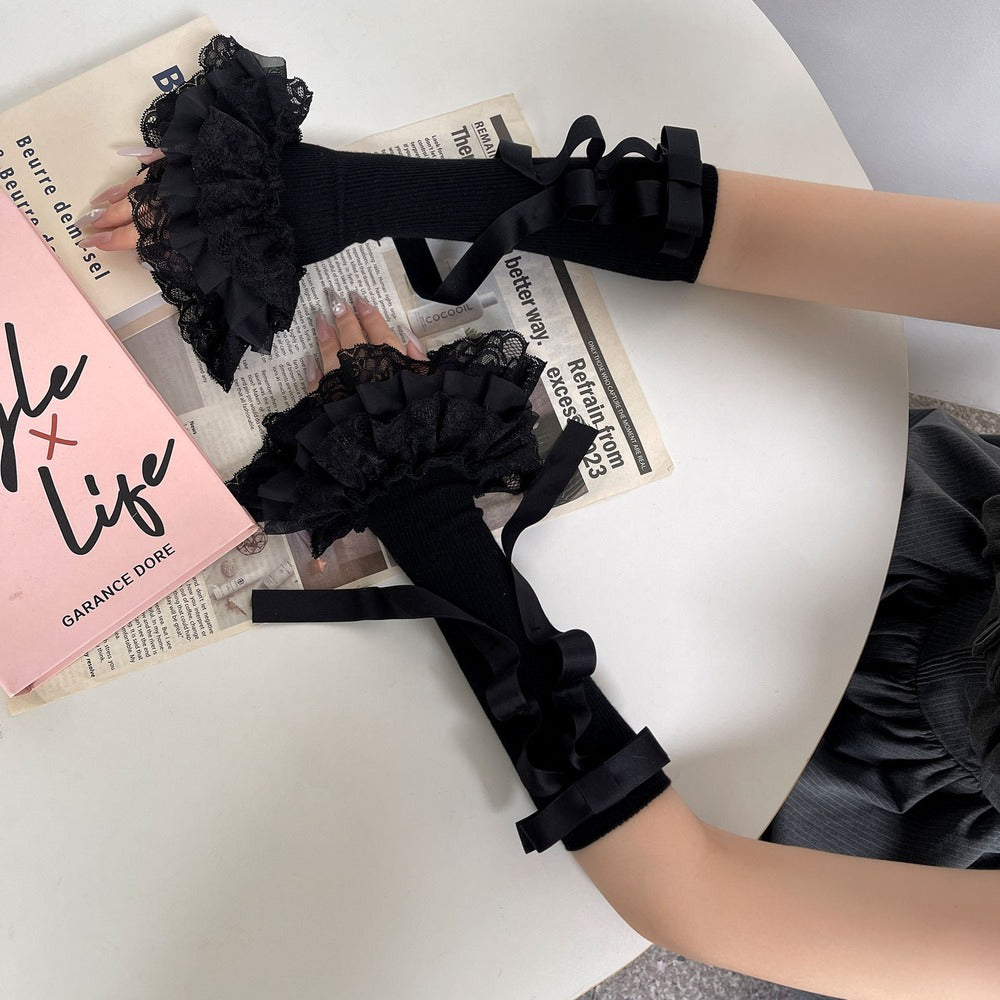 Black lace ruffled long gloves with ribbon bows, shown being worn on a person's hands and arms, styled on top of a newspaper on a white table next to a pink book.