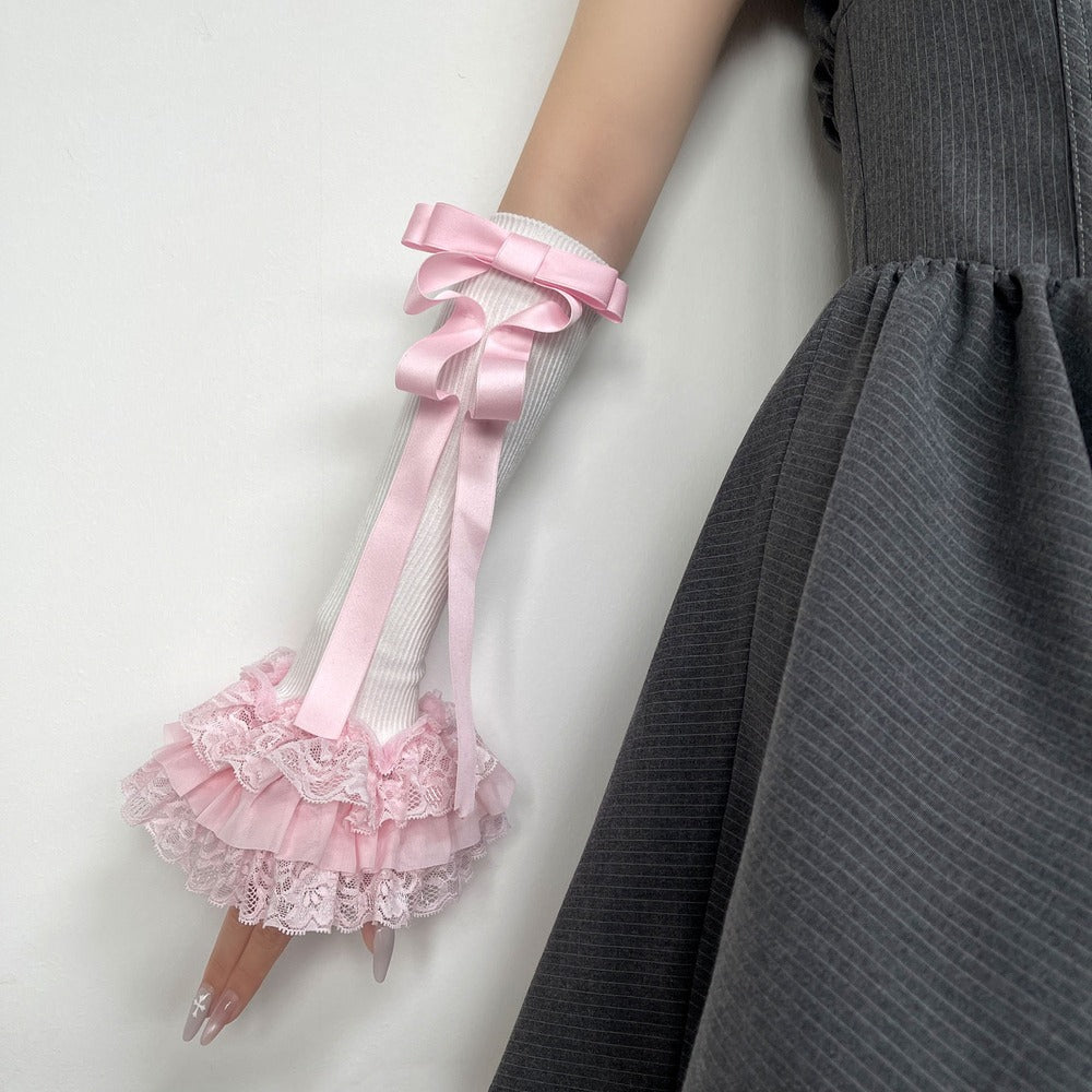 A close-up of a person's arm wearing a long, decorative pink glove with a large satin bow and layered ruffled lace at the cuff, paired with a gray dress against a plain light background.