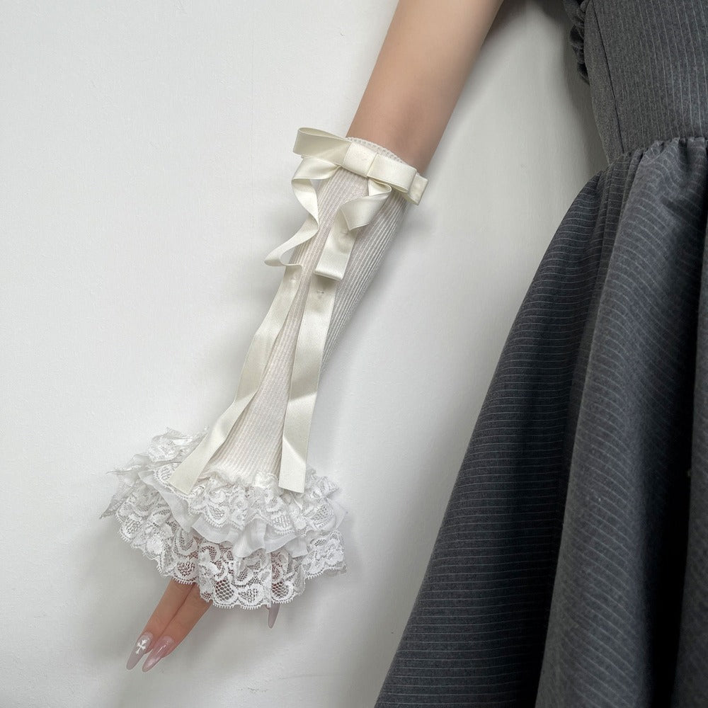 A close-up of a person's hand and forearm wearing a long cream ribbed glove with a large satin bow and a ruffled lace cuff, paired with a dark gray dress.