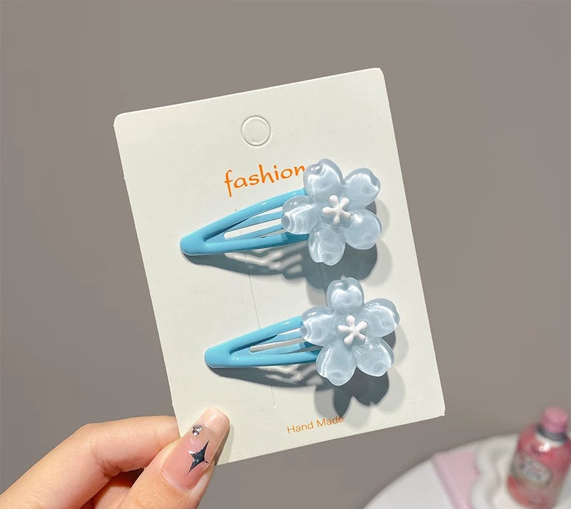 A hand holds a white card displaying two light blue hair clips with translucent pale blue flower charms. The card has the word 'fashion' printed in orange near the top, and the background is a soft gray indoor setting.