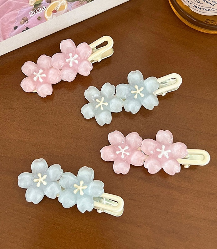 Four pastel flower hair clips arranged on a brown wooden surface. Two clips are light pink with white centers, and two are pale blue with white centers, all with cream-colored metal backs.