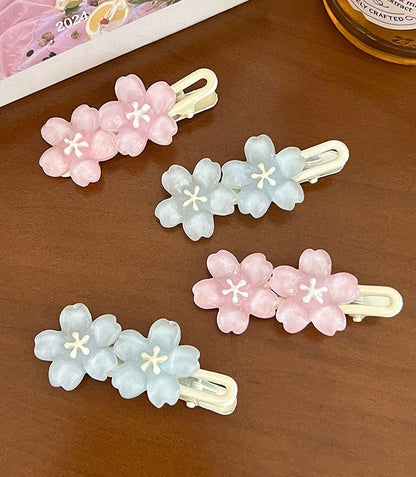 Four pastel flower hair clips arranged on a brown wooden surface. Two clips are light pink with white centers, and two are pale blue with white centers, all with cream-colored metal backs.