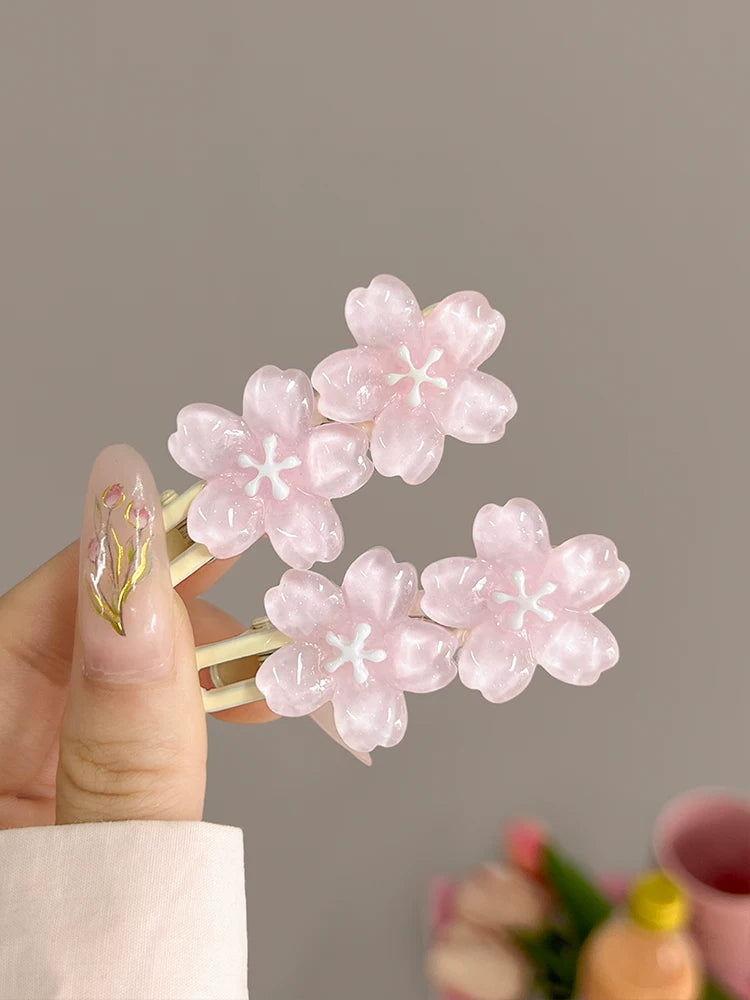 A hand with decorated nails holds a gold-tone hair clip adorned with four glossy light pink cherry blossom flowers against a soft gray background.