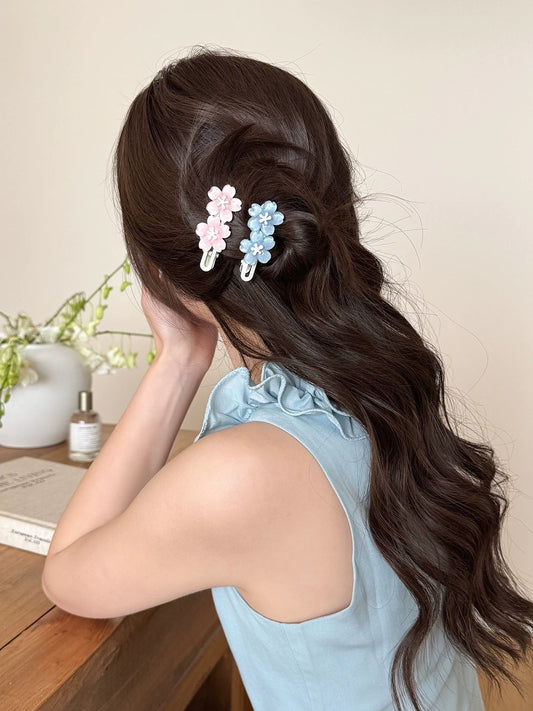 A woman with long, wavy dark brown hair is shown from the back, wearing two decorative hair clips in pastel pink and light blue. She is wearing a light blue sleeveless top and sitting at a wooden table in a softly lit indoor setting.