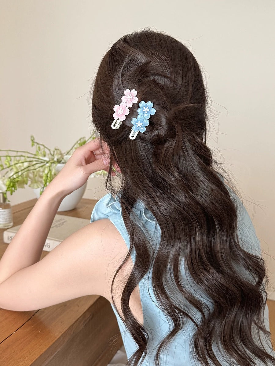 A woman with long, dark brown wavy hair is shown from the back, wearing two small flower-shaped hair clips in pastel pink and light blue. She is wearing a light blue sleeveless top and sitting at a wooden table in a softly lit indoor setting.
